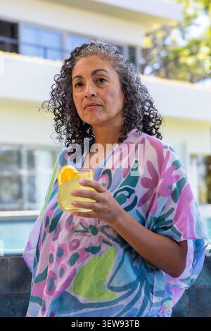 African American woman standing poolside by glass building in coverup holding glass with orange Stock Photo
