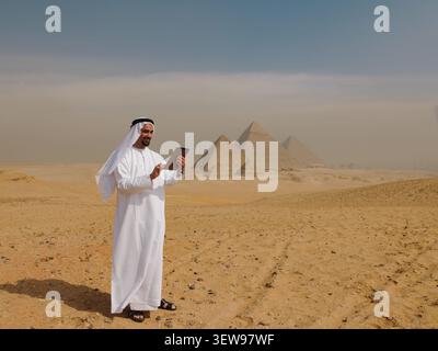 Smiling Arab man in traditional white kandura using a digital tablet in the desert near the Giza pyramids in Egypt ideal for MENA travel tourism and Stock Photo