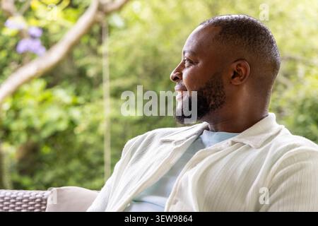 Man sitting on covered porch woven chair, smiling while wearing button-up shirt and tee, copy space Stock Photo