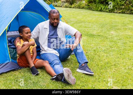 African American father and son sitting by blue tent on grassy lawn, pointing at sleeping bag Stock Photo