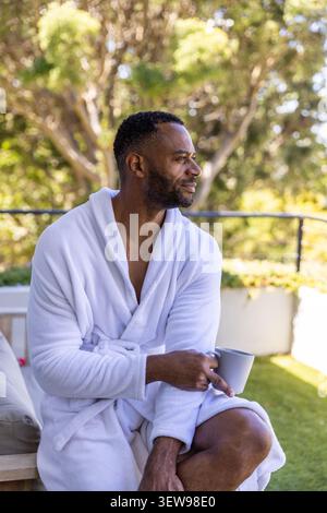 Mature African American man sitting on patio in white robe holding ribbed mug, gazing garden Stock Photo