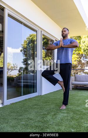 Mature man practicing standing yoga on turf terrace near glass door wearing grey activewear Stock Photo