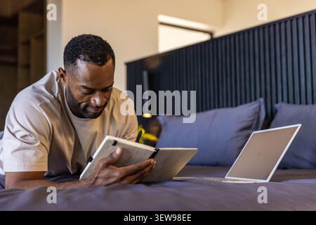 Mature African American man reclining on bed, writing in notebook using pen with laptop, copy space Stock Photo