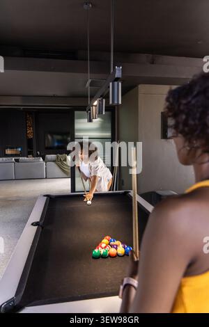 Diverse female friends playing pool in modern game room with black felt table, racked balls, cues Stock Photo