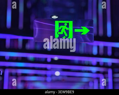 Glowing green emergency exit sign in a futuristic neon-lit interior with vibrant purple and blue lights. Stock Photo