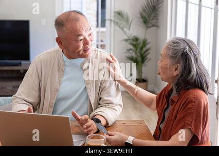 Asian senior couple chatting while using laptop and smartphone at wooden table near windows Stock Photo