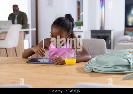 Father and child sitting at wooden table, using stylus on tablet, orange juice, backpack, laptop Stock Photo
