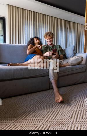 Diverse couple on sofa in living room, woman reclining in orange top, man holding red smartphone Stock Photo