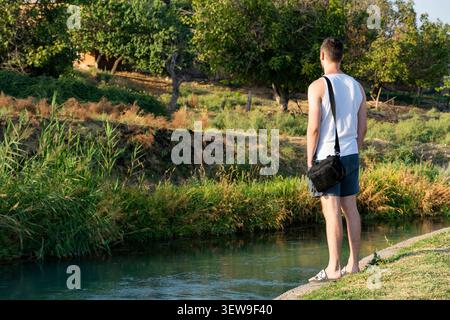 Young man standing on the riverbank surrounded by lush green trees on a warm sunny day. Peaceful outdoor scene representing relaxation, contemplation, Stock Photo