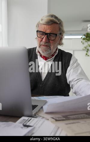 Elderly man sitting at home using a laptop while reviewing household bills, reflecting concern and focus in a quiet domestic setting. Stock Photo