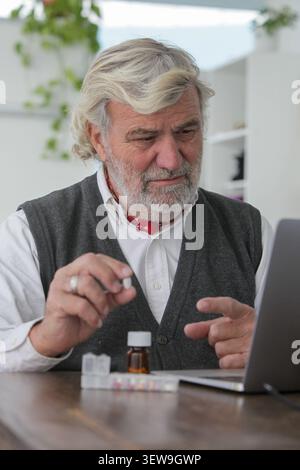 Authentic senior man holding medication while using a laptop during an online consultation at home. Healthcare, telehealth and medication management Stock Photo