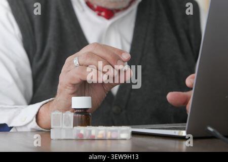 Authentic senior man holding medication while using a laptop during an online consultation at home. Healthcare, telehealth and medication management Stock Photo