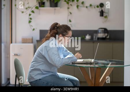 Woman using laptop with poor posture at table feeling neck strain, eye fatigue and screen addiction. Stock Photo