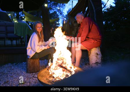Friends relaxing by a campfire at night in a cozy backyard retreat, enjoying conversation and outdoor leisure. Stock Photo