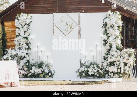 White flowers frame a wedding backdrop with elegant candles. Golden monogram sits above soft, glowing lights. Ceremonial setup invites romance and cel Stock Photo