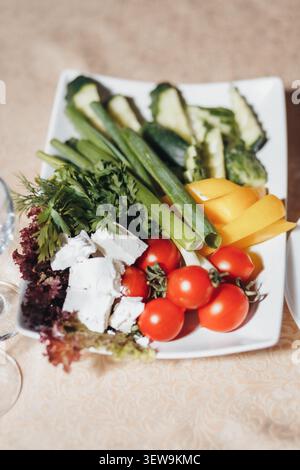 Fresh vegetables and cheese on a white platter. Bright tomatoes, crisp cucumbers, and greens add vibrancy. Platter rests on a soft, warm-toned tablecl Stock Photo
