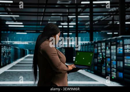 Server room worker uses a green screen laptop in a data center for backend troubleshooting, screen replacement, and server workflow monitoring. Stock Photo