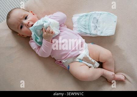 A curious newborn baby lying on a soft beige blanket, holding a fresh disposable diaper near her face and playing with it during changing time. Cute Stock Photo