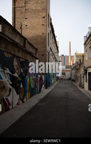 Narrow alley in a former industrial district of Barcelona with brick buildings, colorful street art graffiti on walls, and an old factory chimney Stock Photo