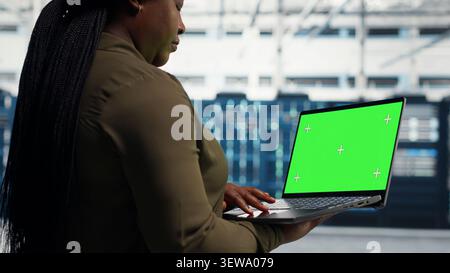 Data center manager uses a green screen mockup computer in a server room for AI workflow and screen replacement. Stock Photo