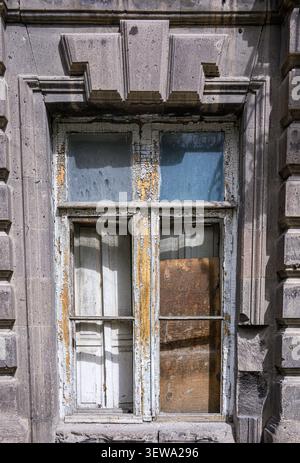 Old weathered window frame with peeling paint and broken glass. Stone wall surrounds worn wooden sash. Sunlight highlights decayed textures. Rust stai Stock Photo