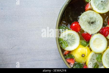 Top view of fruit infused water in metal bowl with lemon slices, strawberries, mint leaves, and ice on rustic gray surface, creating fresh colorful food styling with ample copy space Stock Photo