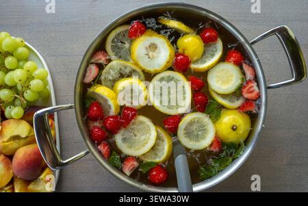 Top view of stainless pot filled with lemon slices, strawberries, mint and ice water beside bowl of green grapes and peaches on wooden table, bright refreshing summer food presentation Stock Photo