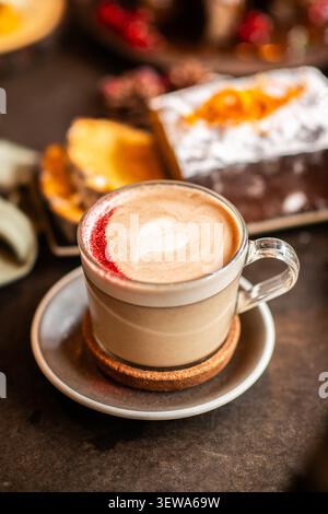 A glass cup of cappuccino featuring heart-shaped latte art and a red powder dusting, served with sliced holiday cake in the background. Stock Photo