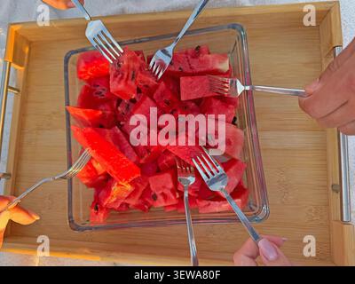 Hands reaching with forks for cubed watermelon in glass dish on wooden tray under  Stock Photo