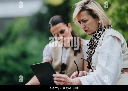 Two professional women collaborating on remote work outdoors with laptop during a focused meeting Stock Photo
