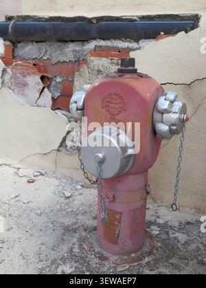 A close-up shot of an old red and pink fire hydrant with silver caps and hanging chains, positioned in front of a cracked, deteriorating stucco wall w Stock Photo