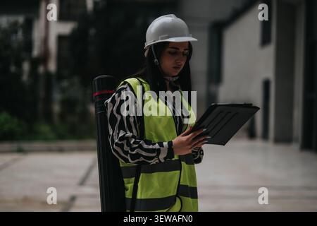 Female engineer on construction site wearing hard hat and high-visibility vest while using a tablet Stock Photo