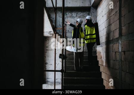 Two construction workers inspecting plans on a concrete stairway at a building site Stock Photo