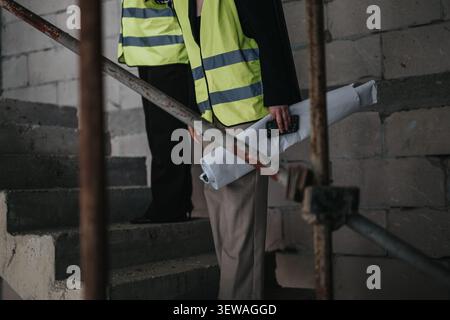 Architect and construction worker inspecting rolled blueprints on stairwell wearing high-visibility safety vests Stock Photo