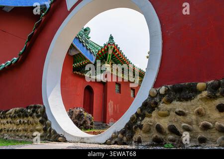 ornate and colourful roof tiles and red walls of koxinga shrine in tainan are framed in white rimmed opening of a red walled moon gate Stock Photo