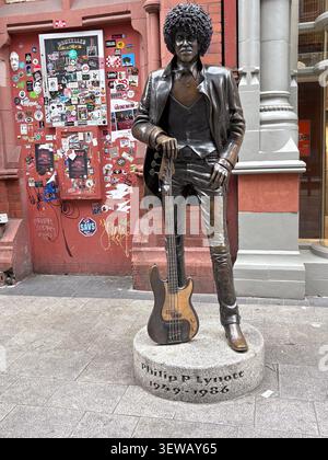 Full-length bronze statue of musician Philip Lynott holding a bass guitar, positioned in front of a red brick wall covered in stickers. Stock Photo