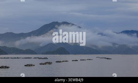 atmospheric seascape of misty mountains rising above a calm coastal sea dotted with floating sea bream fish farms Stock Photo