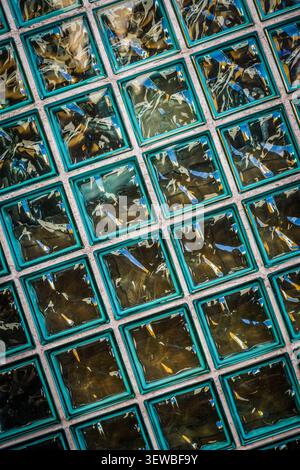 A textured wall composed of square glass blocks arranged in a grid with light gray mortar lines. Each glass block has a raised, wavy, translucent turq Stock Photo