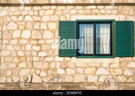 A rustic stone wall features a green window with shutters. The wall is made of various sizes of light-colored stones, creating a textured appearance. Stock Photo