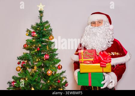 Santa Claus holds boxes with gifts next to a decorated Christmas tree. On the white background. Stock Photo
