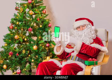 Santa Claus sits in a chair next to the Christmas tree and points to the screen of a smartphone with green screen that he holds in his hand. On the wh Stock Photo