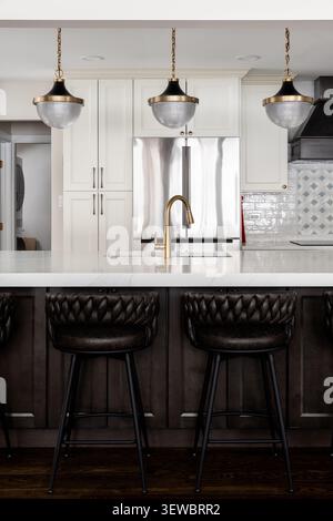 A kitchen detail with black and gold lights hanging over an island, brown cushioned bar stools, cream colored cabinets, and a pattern backsplash. Stock Photo