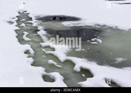 Fresh white snow melting on a frozen river surface during early spring thaw with dark patches of water appearing through thin ice cracks and slushy te Stock Photo