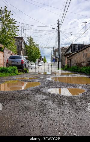 Rainwater pools in deep potholes on a rough urban road. Cars pass along cracked asphalt under power lines. Overhead wires stretch across old buildings Stock Photo
