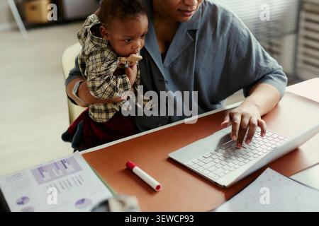 African American mother holding baby while working on laptop in office, balancing childcare with job duties. Ideal for working parent, motherhood, career flexibility marketing Stock Photo