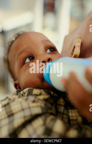Vertical shot of Black infant drinking from baby bottle while resting in mothers arms, showing childcare and feeding routine. Ideal for parenting, maternal care, family support marketing Stock Photo