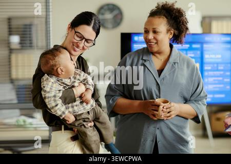 Caucasian young adult mother holding baby while talking with African American female coworker in office, showing working parent support. Ideal for maternity policy or inclusive workplace campaigns Stock Photo