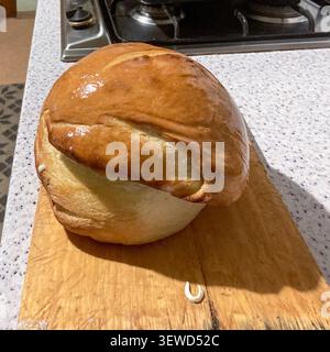 A shiny golden homemade bread loaf rests on a wooden cutting board in a cozy kitchen, with a baking tray and stovetop in the background Stock Photo