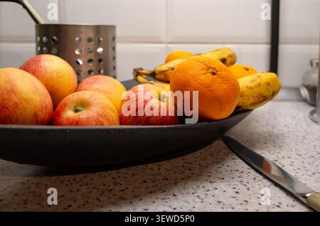 On the speckled kitchen countertop, next to a knife, sits a bowl of fresh fruit: red apples, a bright orange, and ripe bananas Stock Photo