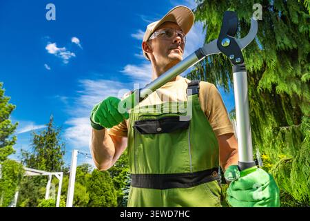 A man stands in a green garden while holding loppers. He wears gloves and is focused on tree maintenance. The sky is clear and blue, with trees visibl Stock Photo